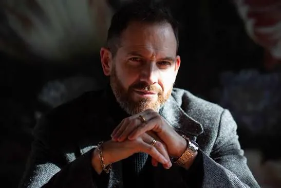 Portrait of author Joe Abercrombie: a bearded man with short hair resting his chin on clasped hands, wearing a grey tweed jacket, lit dramatically against a dark floral backdrop.