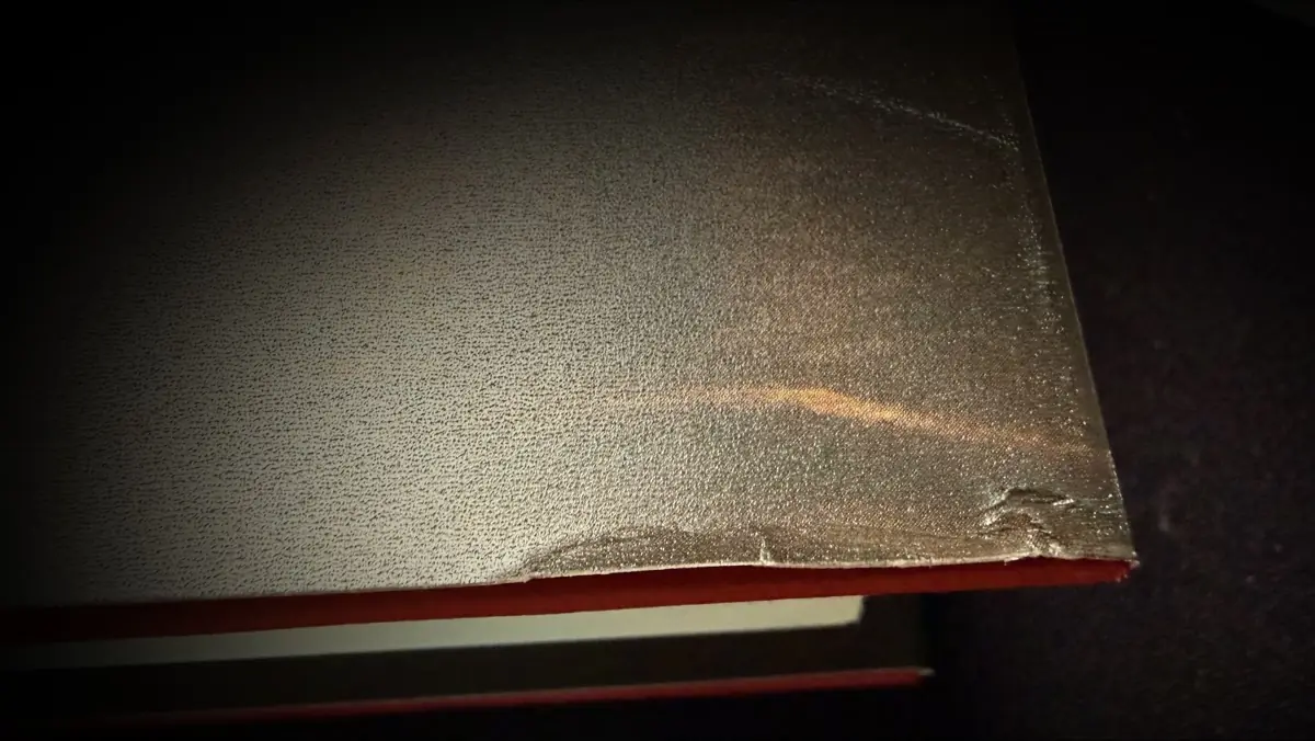 Raking light across the top edge of the Before They Are Hanged dust jacket, showing the gritty finish scuffed pale and the corner of the jacket torn and lifted away from the red board underneath.