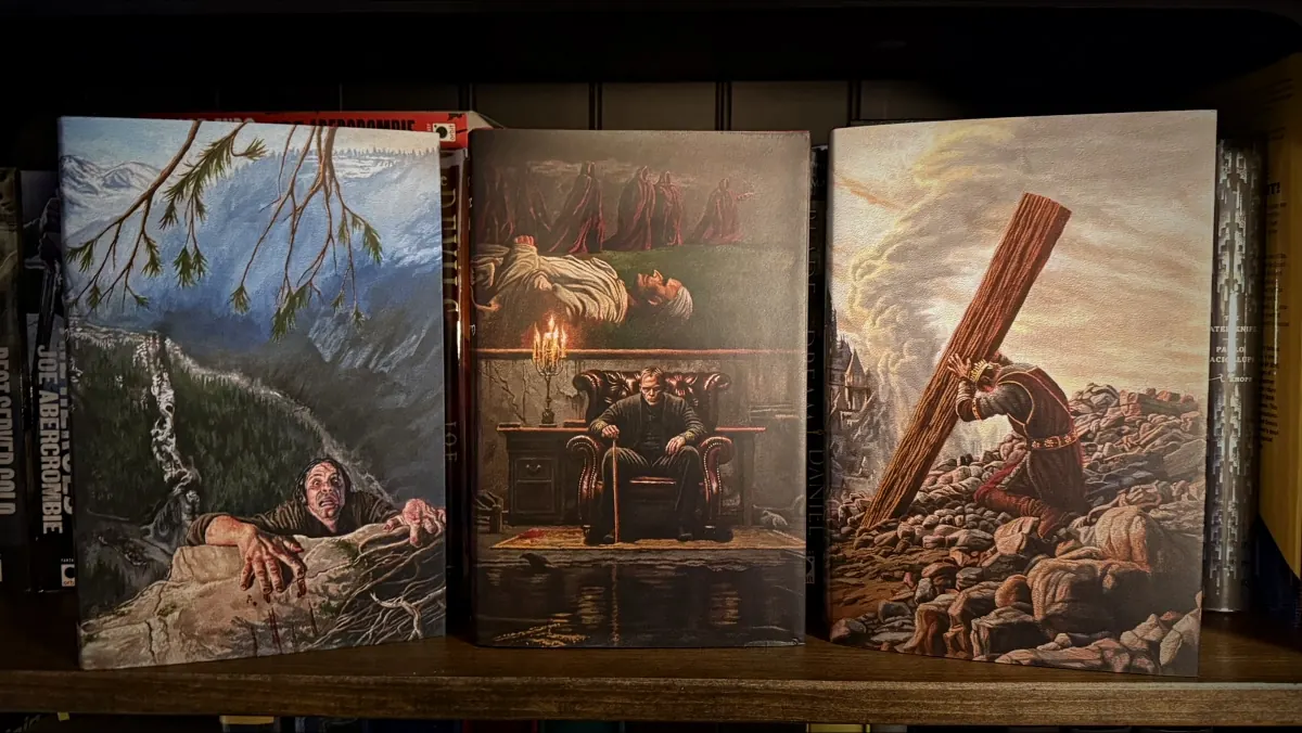 The three Lit Escalates First Law dust jackets standing upright on a shelf, showing Joel Daniel Phillips oil paintings: Logen Ninefingers clinging to a cliff above a river, Bayaz seated in a chair before a candlelit desk with hooded figures above, and an armoured figure hauling a wooden beam through rubble.