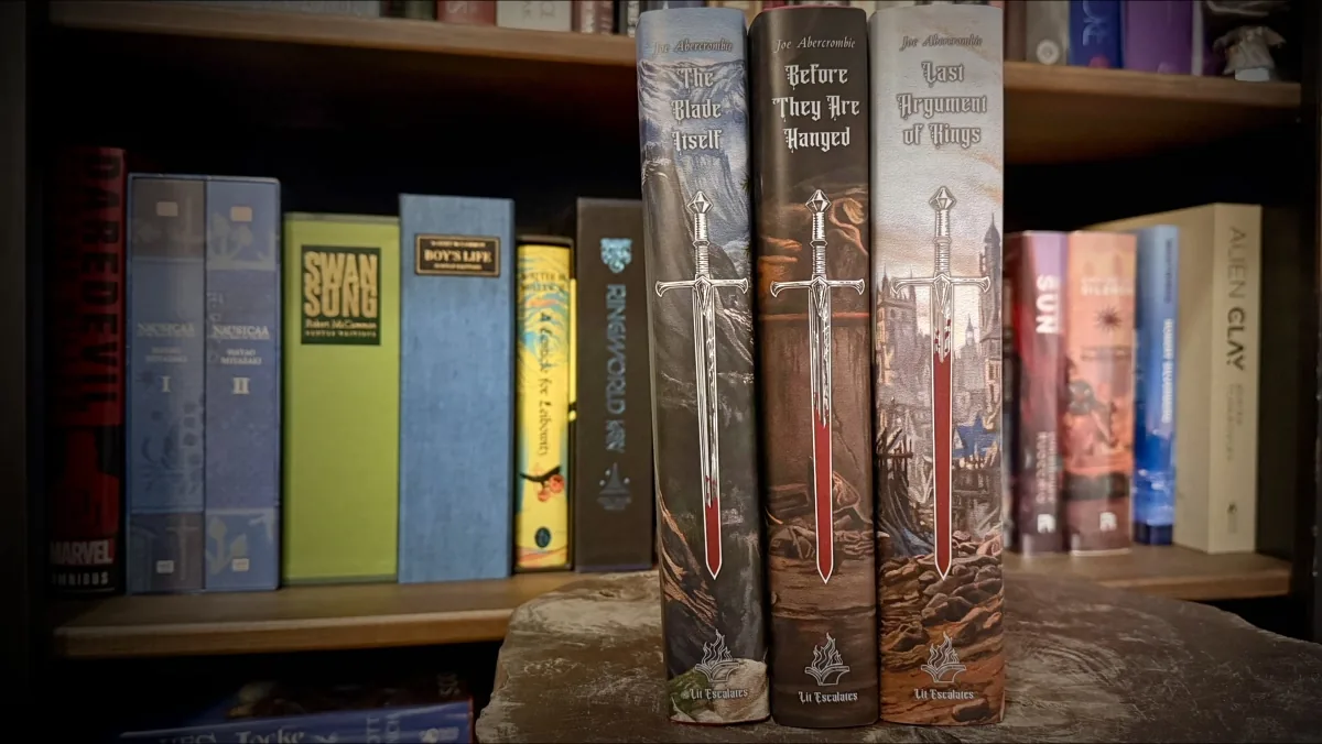 The spines of the three Lit Escalates First Law hardcovers side by side, each printed with a sword. Blood covers the lowest portion of the blade on The Blade Itself, rises higher on Before They Are Hanged, and reaches highest on Last Argument of Kings, with the Lit Escalates logo at the foot of each spine.
